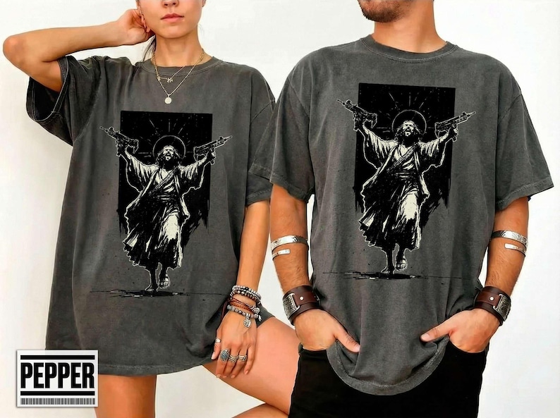 May include: A charcoal gray short-sleeve t-shirt featuring a black and white graphic print of a figure in robes with a halo, holding a firearm in each outstretched hand. The design is centered on the chest of the garment.