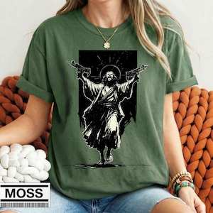 May include: An olive green t-shirt featuring a black and white graphic print of a figure with a halo holding two firearms. The figure is depicted in a robe with a bandolier across the chest. The shirt has short sleeves and a crew neckline.