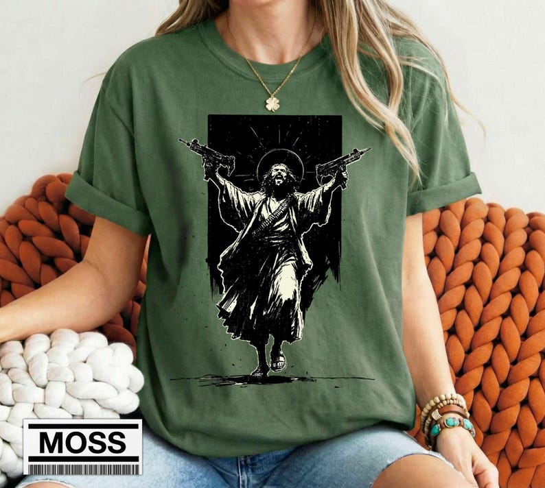 May include: An olive green t-shirt featuring a black and white graphic print of a figure with a halo holding two firearms. The figure is depicted in a robe with a bandolier across the chest. The shirt has short sleeves and a crew neckline.
