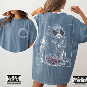 May include: A blue jean colored t-shirt featuring a front chest logo that reads THE DESPERADO CLUB with a dagger graphic. The back displays a large white line art illustration of a cat wearing a crown and sunglasses next to a bowl of cherries.