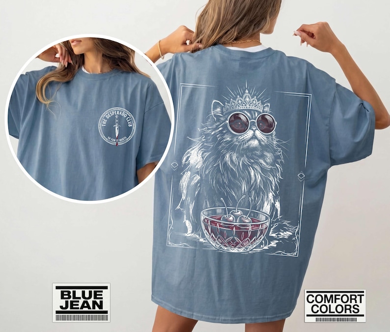 May include: A blue jean colored t-shirt featuring a front chest logo that reads THE DESPERADO CLUB with a dagger graphic. The back displays a large white line art illustration of a cat wearing a crown and sunglasses next to a bowl of cherries.