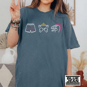May include: A slate blue short-sleeve t-shirt featuring a white graphic print of boxer shorts with red hearts, a cat wearing a gold crown and round glasses, and a dinosaur head with a pink crest. The shirt has a crew neckline and a relaxed fit.