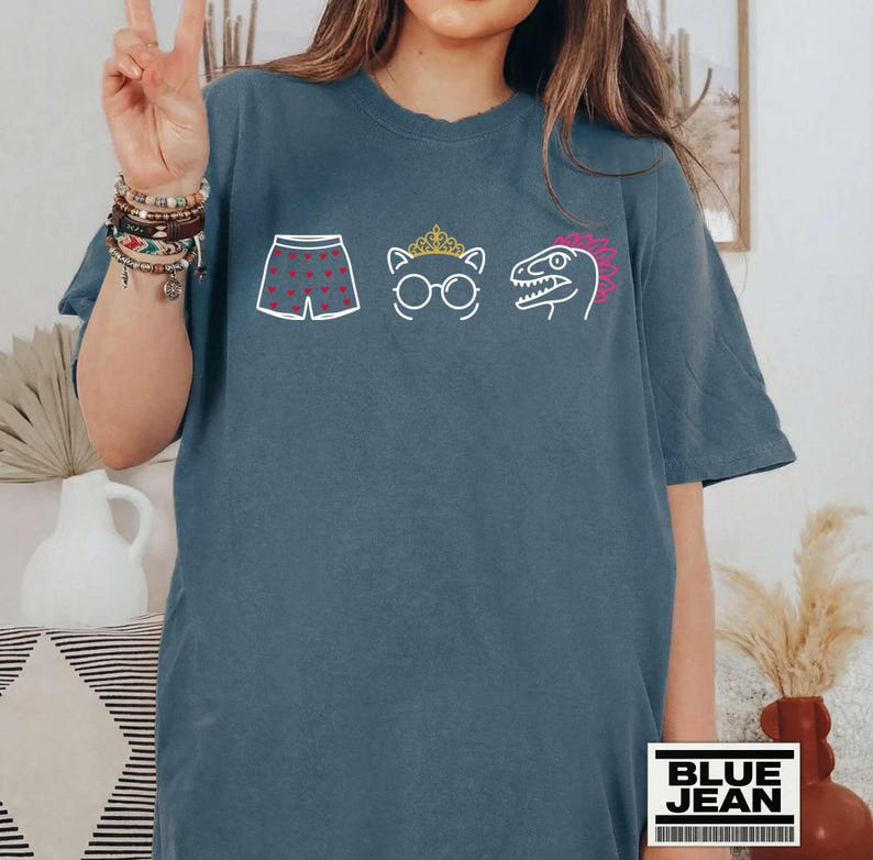 May include: A slate blue short-sleeve t-shirt featuring a white graphic print of boxer shorts with red hearts, a cat wearing a gold crown and round glasses, and a dinosaur head with a pink crest. The shirt has a crew neckline and a relaxed fit.