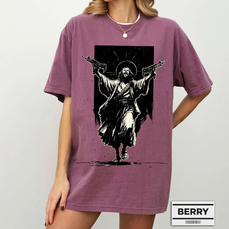 May include: A berry-colored short-sleeve t-shirt featuring a black and white graphic print of a figure with long hair and a beard, wearing robes, holding a firearm in each hand, and standing against a dark rectangular background. The text BERRY is visible.