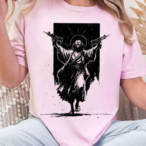 May include: A light pink short-sleeved t-shirt featuring a black graphic print of a figure with long hair and a halo, wearing robes and holding two firearms aloft. The design is set against a solid black rectangular background on the chest.