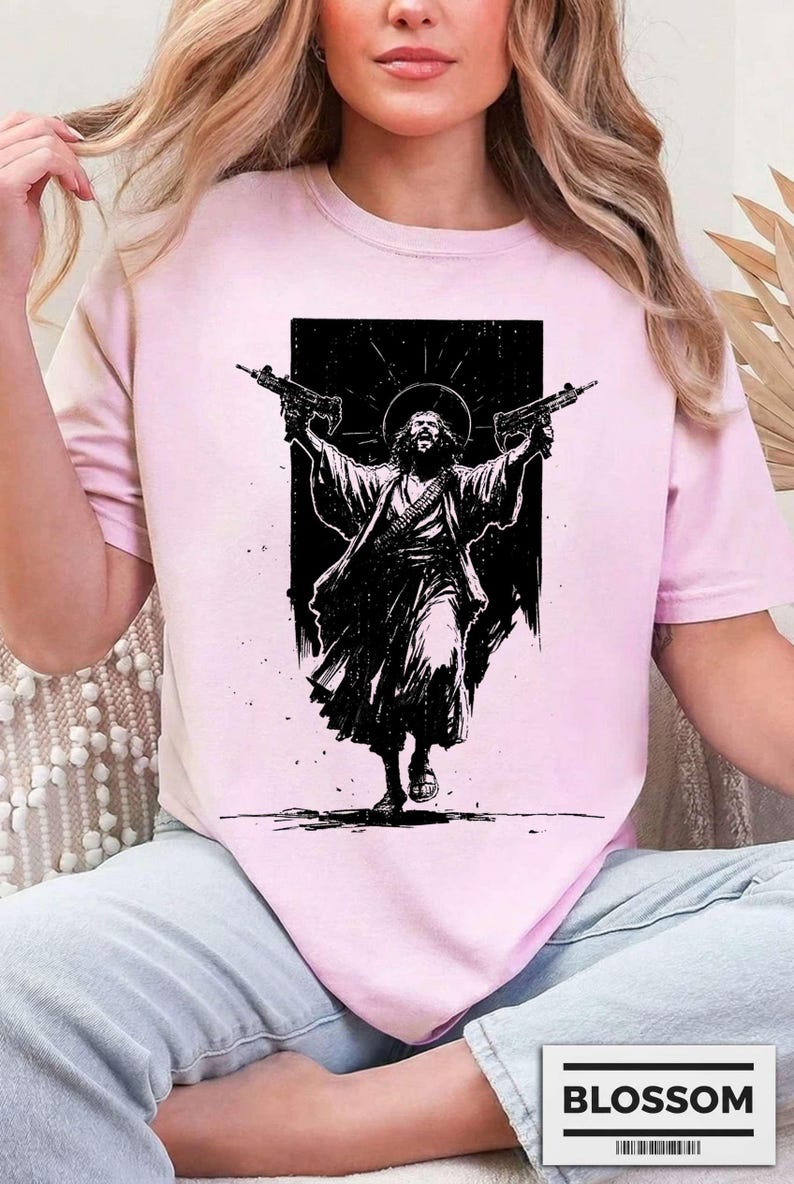 May include: A light pink short-sleeved t-shirt featuring a black graphic print of a figure with long hair and a halo, wearing robes and holding two firearms aloft. The design is set against a solid black rectangular background on the chest.