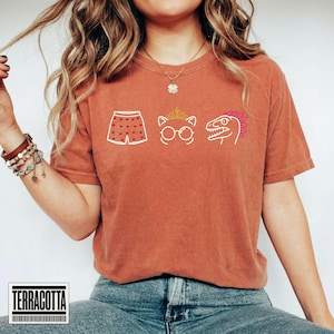 May include: A terracotta-colored short-sleeve t-shirt featuring a graphic print of white and gold line art icons. The icons include a pair of shorts with a heart pattern, a cat wearing a crown and glasses, and a dinosaur head with a pink crest.