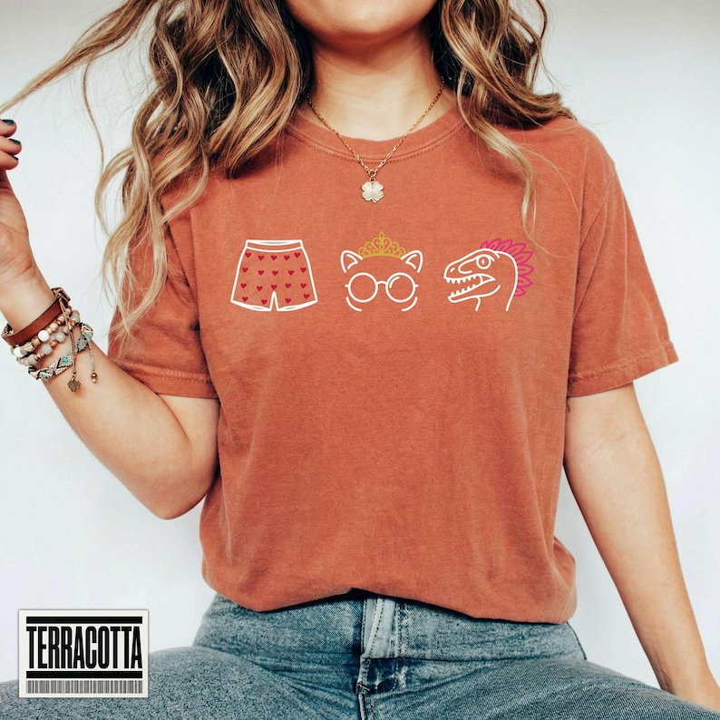 May include: A terracotta-colored short-sleeve t-shirt featuring a graphic print of white and gold line art icons. The icons include a pair of shorts with a heart pattern, a cat wearing a crown and glasses, and a dinosaur head with a pink crest.