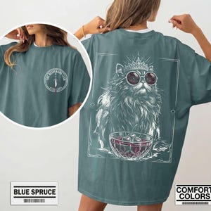 May include: A blue spruce t-shirt featuring a front chest logo with a dagger and the text The Desperado Club. The back graphic displays a long-haired cat wearing a crown and round sunglasses, sitting behind a bowl of cherries in a square frame.