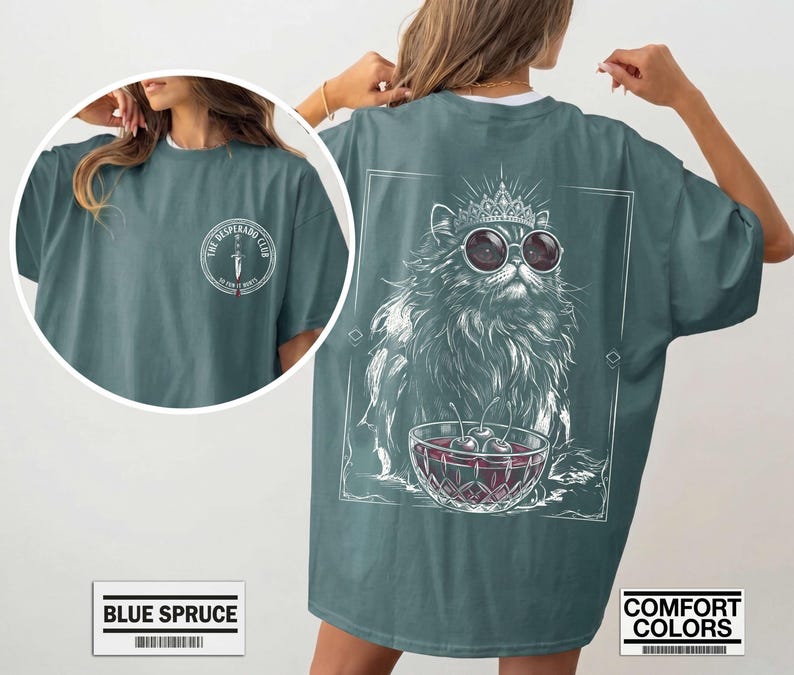 May include: A blue spruce t-shirt featuring a front chest logo with a dagger and the text The Desperado Club. The back graphic displays a long-haired cat wearing a crown and round sunglasses, sitting behind a bowl of cherries in a square frame.