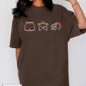 May include: A dark brown short-sleeve t-shirt featuring a graphic print of white boxer shorts with red hearts, a white cat outline wearing a gold crown and glasses, and a white dinosaur head with a pink crest. The word ESPRESSO is printed below.