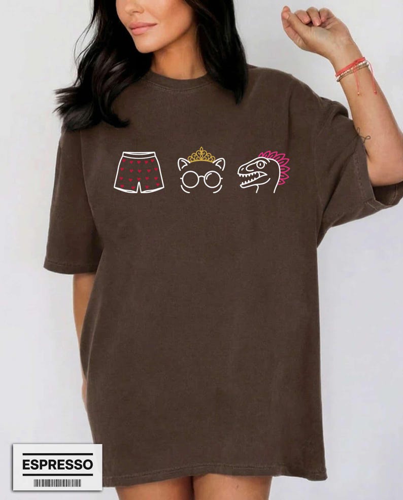 May include: A dark brown short-sleeve t-shirt featuring a graphic print of white boxer shorts with red hearts, a white cat outline wearing a gold crown and glasses, and a white dinosaur head with a pink crest. The word ESPRESSO is printed below.