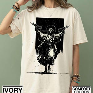 May include: An ivory-colored short-sleeve t-shirt featuring a black graphic print of a figure with a halo, wearing robes and holding two submachine guns aloft. The design is centered on the chest, showcasing a high-contrast, ink-style illustration.