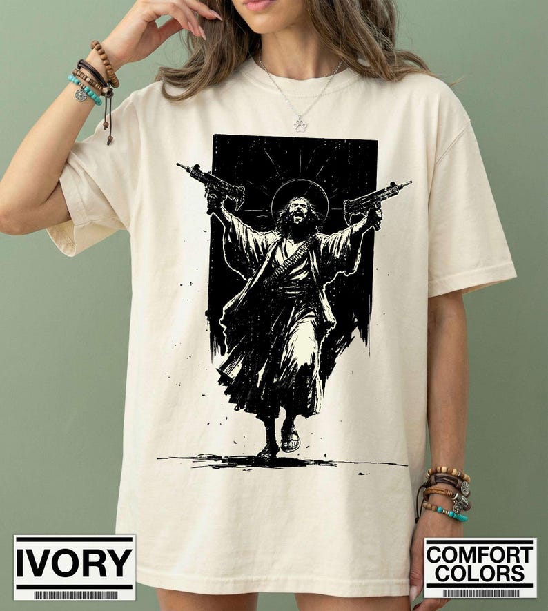 May include: An ivory-colored short-sleeve t-shirt featuring a black graphic print of a figure with a halo, wearing robes and holding two submachine guns aloft. The design is centered on the chest, showcasing a high-contrast, ink-style illustration.