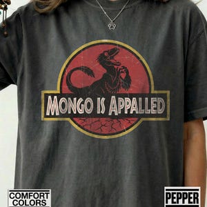 May include: A charcoal gray short-sleeve t-shirt featuring a circular graphic with a red background and a black dinosaur silhouette. The text "MONGO IS APPALED" is printed in white capital letters across a black rectangular banner in the center.