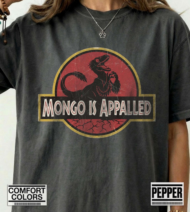 Może przedstawiać: Ciemnoszary t-shirt z kr&oacute;tkim rękawem z okrągłą grafiką na czerwonym tle, przedstawiającą czarną sylwetkę dinozaura. Napis "MONGO IS APPALED" jest wydrukowany białymi wielkimi literami na czarnym prostokątnym banerze umieszczonym pośrodku.