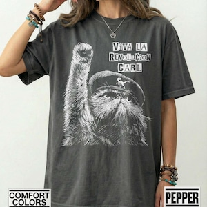 May include: Dark gray t-shirt featuring a graphic of a cat wearing a beret and raising a paw, with the text "VIVA LA REVOLUTION CARL" in white. The shirt also has the text "COMFORT COLORS" and "PEPPER" in white.