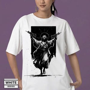 May include: A white short-sleeved t-shirt featuring a black graphic print of a bearded figure with a halo, wearing robes and holding two submachine guns aloft. The design is centered on the chest, with the word WHITE printed in a small box below.