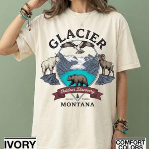 May include: Cream-coloured t-shirt with a graphic featuring the word "GLACIER" above a circular design. The design includes an eagle, mountain goats, a bear, and the text "Outdoor Discovery" and "Montana". The shirt is labelled "Comfort Colors".