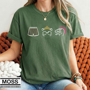 May include: An olive green short-sleeved t-shirt featuring a white graphic print of boxer shorts with red hearts, a cat wearing a crown and round glasses, and a dinosaur head with a pink crest. The shirt has a crew neckline and rolled sleeves.
