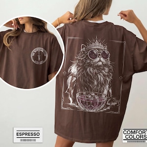 May include: A brown t-shirt featuring a white graphic on the back of a fluffy cat wearing a crown and round sunglasses, sitting behind a bowl of cherries. The front chest area displays a circular logo with a dagger and the text THE DESPERADO CLUB.