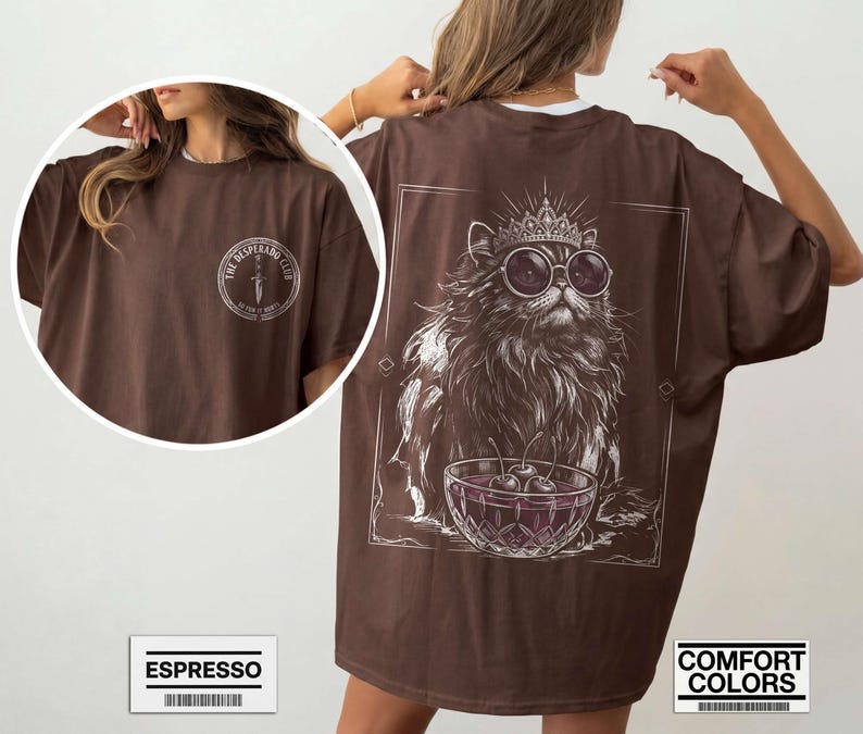 May include: A brown t-shirt featuring a white graphic on the back of a fluffy cat wearing a crown and round sunglasses, sitting behind a bowl of cherries. The front chest area displays a circular logo with a dagger and the text THE DESPERADO CLUB.