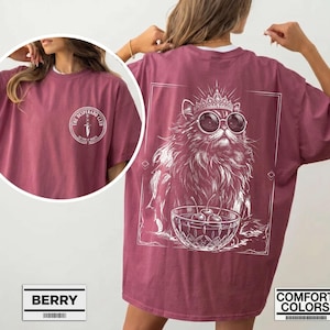 May include: A berry-colored t-shirt featuring a white graphic on the back of a fluffy cat wearing a crown and sunglasses next to a bowl of cherries. The front chest area displays a circular logo with a dagger and the text The Desperado Club.