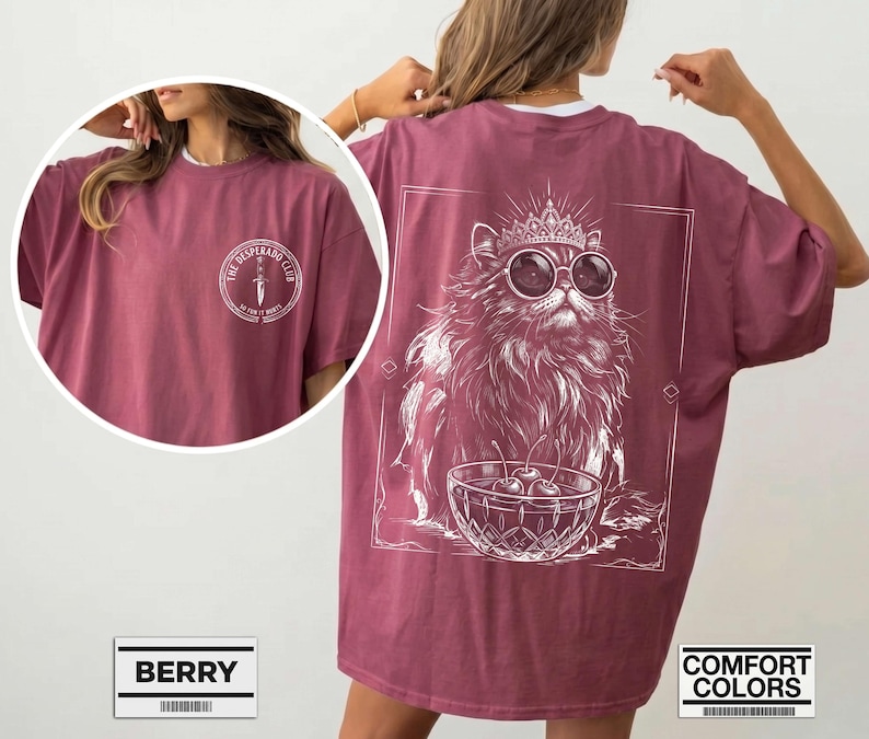 May include: A berry-colored t-shirt featuring a white graphic on the back of a fluffy cat wearing a crown and sunglasses next to a bowl of cherries. The front chest area displays a circular logo with a dagger and the text The Desperado Club.