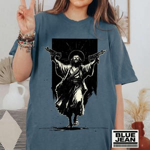 May include: A slate blue t-shirt featuring a black and white graphic print of a figure with a halo holding two firearms. The design depicts a robed individual with long hair and a beard, arms raised, set against a dark rectangular background.