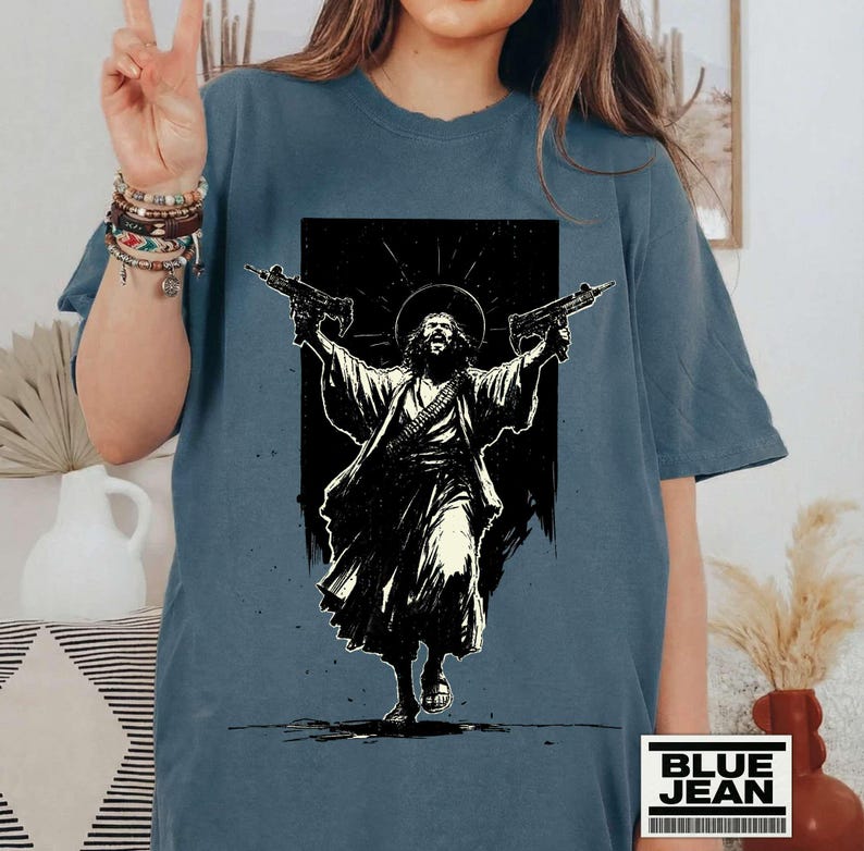 May include: A slate blue t-shirt featuring a black and white graphic print of a figure with a halo holding two firearms. The design depicts a robed individual with long hair and a beard, arms raised, set against a dark rectangular background.