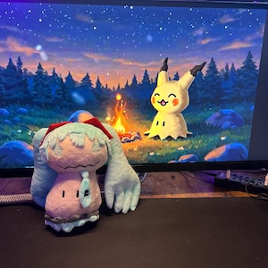 May include: A plush toy of a light blue and pink character with large arms and a red hat, and a yellow Pikachu plush toy are on a desk in front of a computer screen displaying a campfire scene. The screen also shows the text "Privacy Report a bug | Flag as objectionable" and "under Creative Commons Attribution-ShareAlike 3.0 License".