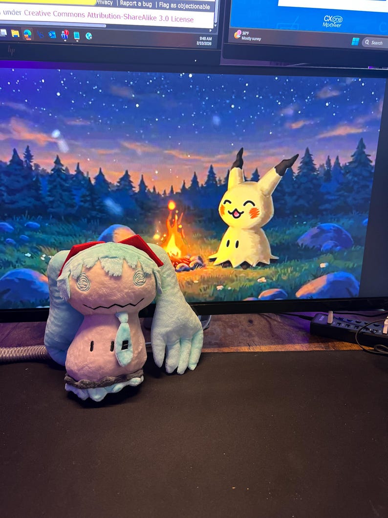 May include: A plush toy of a light blue and pink character with large arms and a red hat, and a yellow Pikachu plush toy are on a desk in front of a computer screen displaying a campfire scene. The screen also shows the text "Privacy Report a bug | Flag as objectionable" and "under Creative Commons Attribution-ShareAlike 3.0 License".