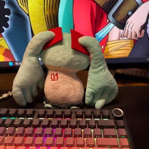 May include: A plush toy with a light blue body, red head pieces, and large hands. The toy has the number "01" printed on its front. It sits in front of a backlit keyboard with pink and purple keys.