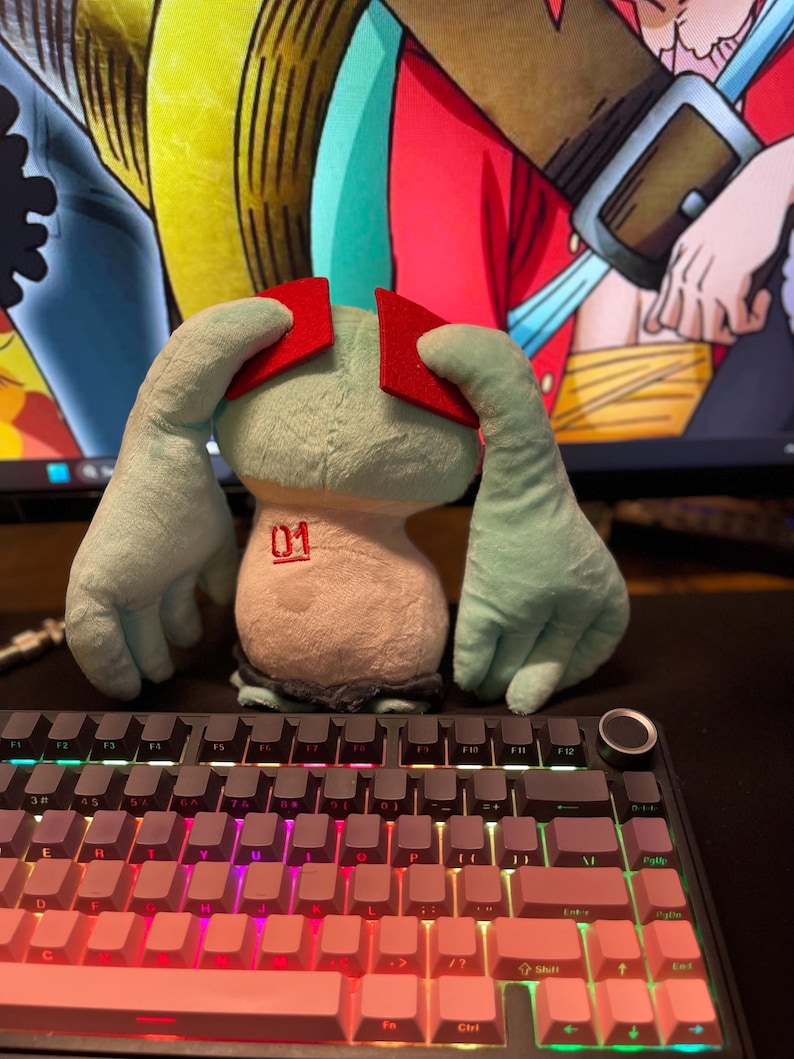 May include: A plush toy with a light blue body, red head pieces, and large hands. The toy has the number "01" printed on its front. It sits in front of a backlit keyboard with pink and purple keys.