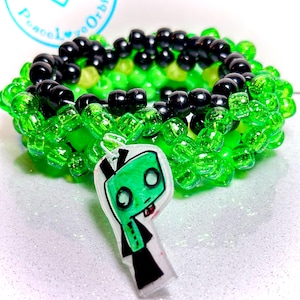 May include: A kandi bracelet featuring black, green, and yellow beads. A clear charm with a green cartoon character is attached. The bracelet is a vibrant accessory, ideal for raves or festivals.