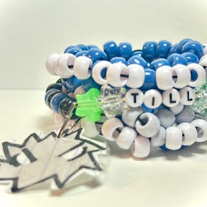 May include: A stack of blue and white beaded bracelets with the word "TILL" spelled out in white beads. A clear charm with a black and white graphic is attached to the bracelet.