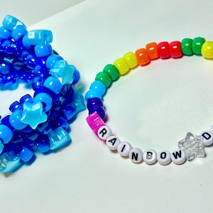 May include: Two beaded bracelets. One is a cuff made of blue beads and star-shaped accents. The other is a rainbow-colored bracelet with the words "RAINBOW DASH" spelled out in white letter beads, with star-shaped accents.