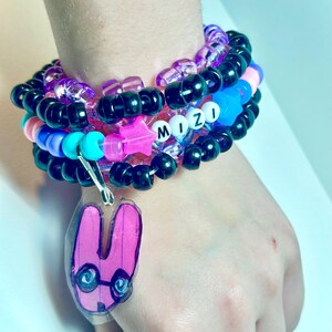 Puede incluir: Una pila de pulseras de cuentas en varios colores, incluyendo negro, morado, rosa, azul y turquesa. Las pulseras tienen las letras "MIZI" deletreadas en cuentas blancas. Un dije de conejo rosa con gafas cuelga de las pulseras.