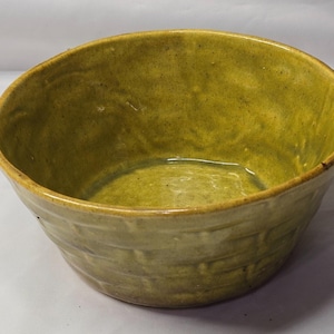 Puede incluir: Un cuenco de cerámica de forma redonda, moldeado a mano, con un esmalte brillante de color amarillo mostaza. El exterior presenta una textura de tejido de cesta, mientras que el interior es liso con sutiles variaciones de color.