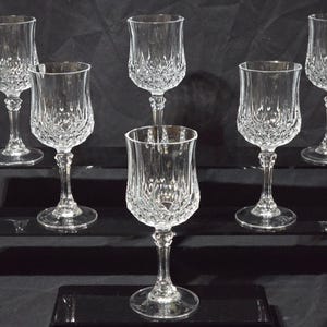 Può includere: Un set di sei calici in vetro trasparente caratterizzati da un motivo a sfaccettature a diamante sulla parte inferiore della coppa e da uno stelo sottile con un nodo decorativo. Ogni calice presenta un bordo liscio e una base circolare.