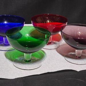 May include: A set of four glass dessert bowls featuring a wide, shallow basin and a short, clear stem with a circular base. The bowls are colored in vibrant shades of cobalt blue, emerald green, deep ruby red, and a rich, dark purple hue.