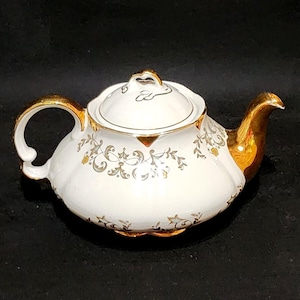 Puede incluir: Una tetera de porcelana blanca con asa, pico y remate de tapa pintados en oro. El cuerpo está decorado con un delicado patrón de volutas florales en gris y oro. La tetera presenta un diseño de bordes festoneados y una forma redondeada.
