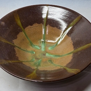 Può includere: Una ciotola in ceramica larga e poco profonda con un bordo esterno marrone scuro che sfuma verso un centro color sabbia più chiaro. La superficie interna è decorata con gocce di smalto verde radianti che partono dal centro verso il bordo.