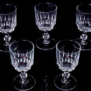 Peut inclure: Un ensemble de cinq verres à pied en verre transparent présentant un motif côtelé vertical sur le bas du calice et une tige à facettes. Chaque verre possède une base circulaire et un calice cylindrique, disposés sur un fond sombre.