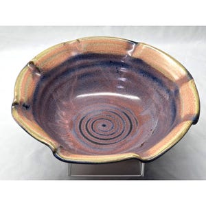 May include: A ceramic bowl with a scalloped rim, featuring a glaze of blended blue, purple, and tan tones. The interior base displays a dark, concentric circular pattern, while the overall surface has a smooth, glossy finish throughout.