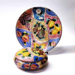 Puede incluir: Un plato decorativo y un recipiente redondo a juego con tapa, que presentan un colorido patrón de mosaico de varias frutas, incluyendo limones, uvas y granadas. El diseño utiliza pequeñas piezas texturizadas en tonos azul, rosa, amarillo y verde.