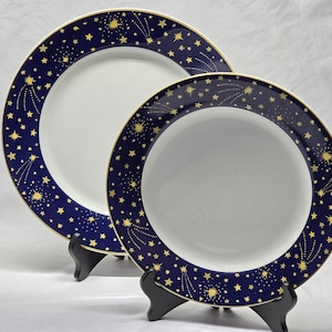 Puede incluir: Dos platos redondos de cerámica con centro blanco y un borde ancho de color azul oscuro. El borde presenta un patrón de color dorado con estrellas de varios tamaños y estrellas fugaces. Cada plato tiene un borde fino dorado en los extremos.
