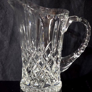 May include: A clear glass pitcher featuring a diamond-cut pattern on the lower body and vertical fluted panels on the upper section. The vessel includes a curved handle with a textured grip and a wide pouring spout at the top rim.