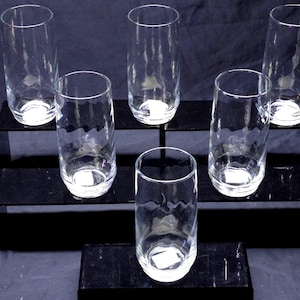 May include: A set of five clear glass tumblers with a cylindrical shape and a subtle textured pattern on the exterior. Each glass features a smooth rim and a thick, weighted base, arranged in a tiered formation to display the transparent design.