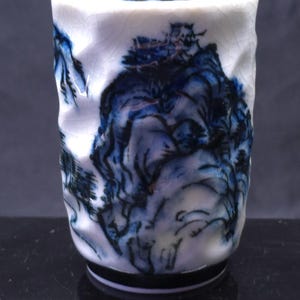 May include: A white ceramic cup with a textured, uneven surface featuring a hand-painted blue and black ink wash landscape design. The cup has a cylindrical shape with a crackled glaze finish and a solid black base ring at the bottom edge.